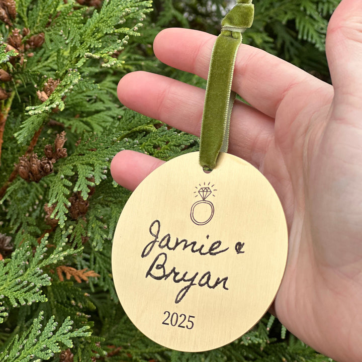 Hand holding a gold ornament tag with 'Jamie & Bryan 2025' text against a green Christmas tree background.